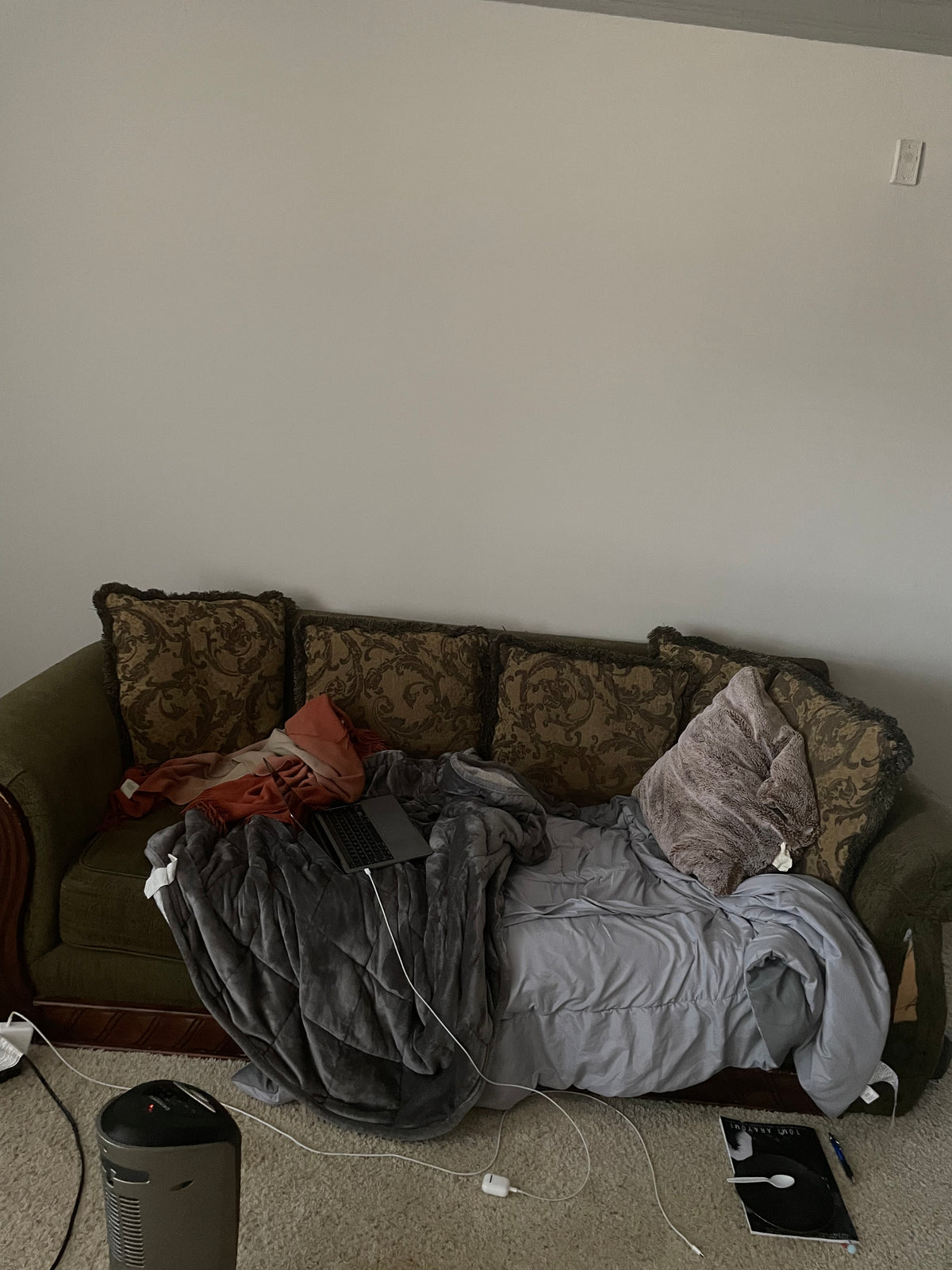 A cluttered olive green couch in a living room setting, covered with a gray blanket, patterned throw pillows, and an orange pillow, with a laptop and various cables on the floor.