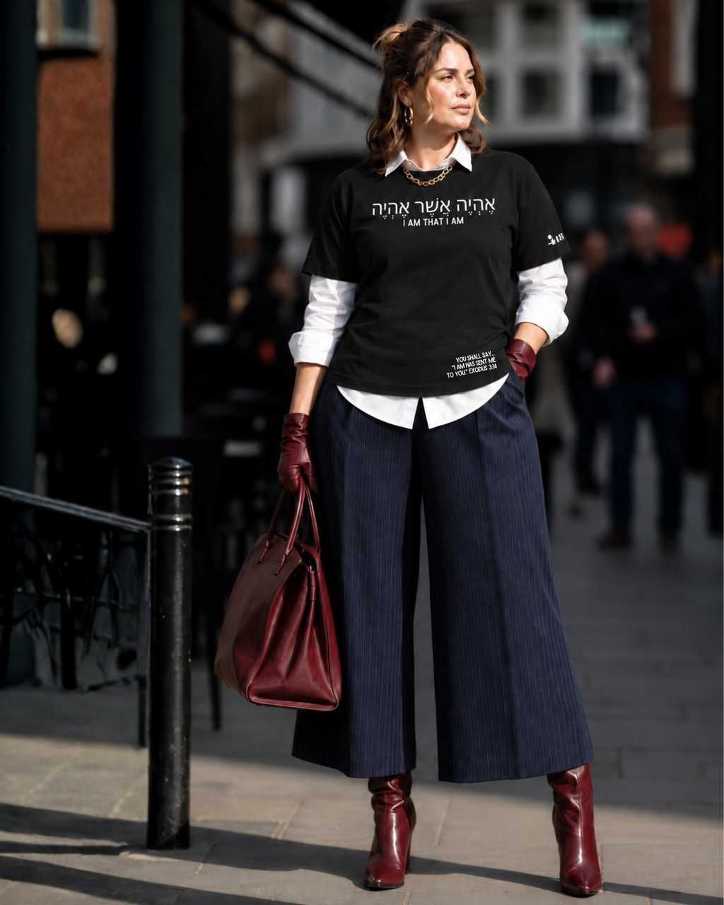A woman stands on a city street wearing a Project 418 Co black graphic T-shirt with white text,  ('I AM THAT I AM') and additional small white text near the lower hem, layered over a white collared shirt, dark wide-leg pinstripe trousers, burgundy gloves, and burgundy ankle boots. She carries a large burgundy handbag.