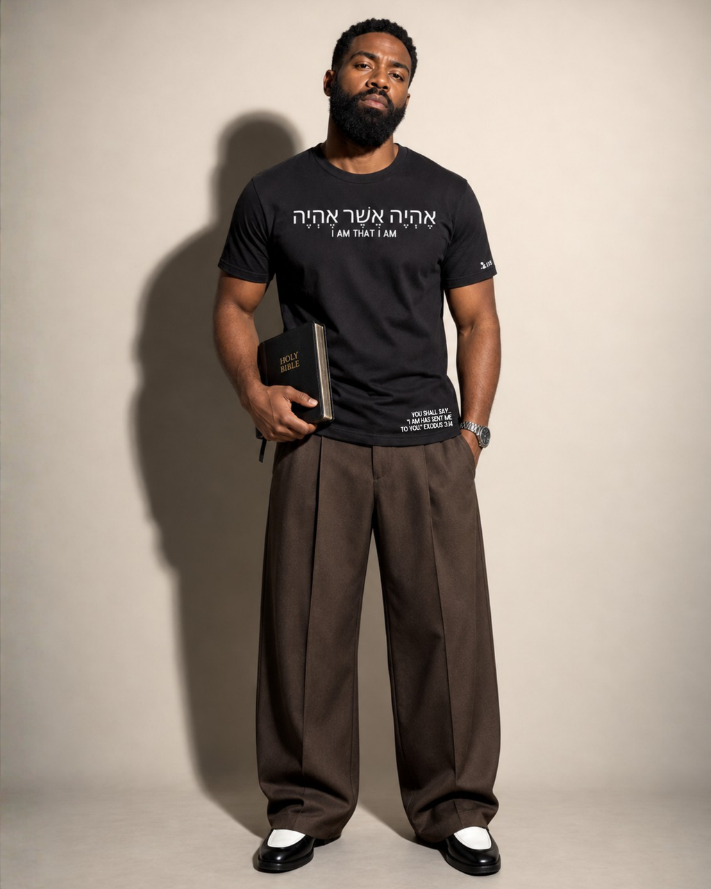 A man stands against a plain beige studio background wearing a Project 418 Co black graphic T-shirt with white Hebrew and English text,  ('I AM THAT I AM') and additional small white text near the lower hem, loose brown trousers, and black-and-white shoes. He holds a dark book or notebook in one hand.