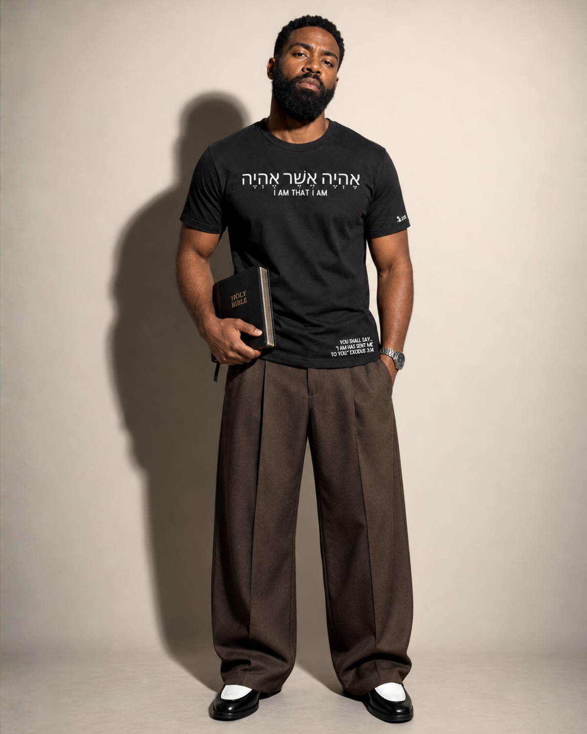 A man stands against a plain beige studio background wearing a Project 418 Co black graphic T-shirt with white Hebrew and English text,  ('I AM THAT I AM') and additional small white text near the lower hem, loose brown trousers, and black-and-white shoes. He holds a dark book or notebook in one hand.