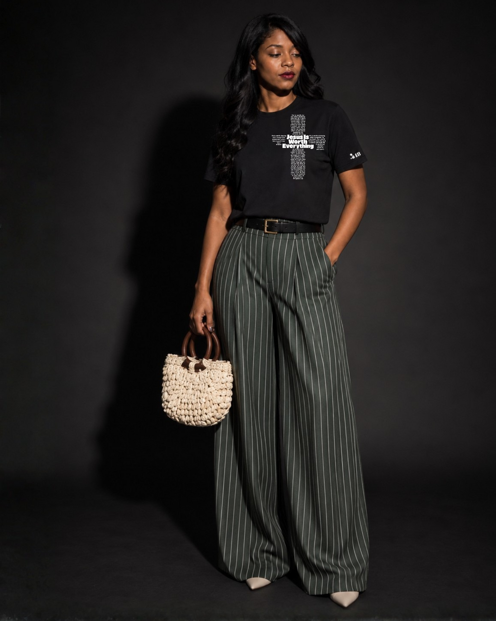 A woman stands against a dark studio background wearing a Project 418 Co black graphic T-shirt with white design writing of Jesus is Everything in a cross form, wide-leg dark green pinstriped trousers, a black belt, and holding a small woven cream handbag.