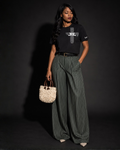 A woman stands against a dark studio background wearing a Project 418 Co black graphic T-shirt with white design writing of Jesus is Everything in a cross form, wide-leg dark green pinstriped trousers, a black belt, and holding a small woven cream handbag.
