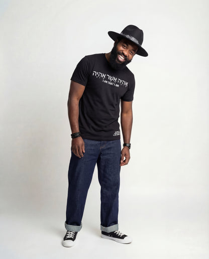 Man wearing a black Mission Tee "I Am" with hebrew text, dark jeans, and a black hat on a white background