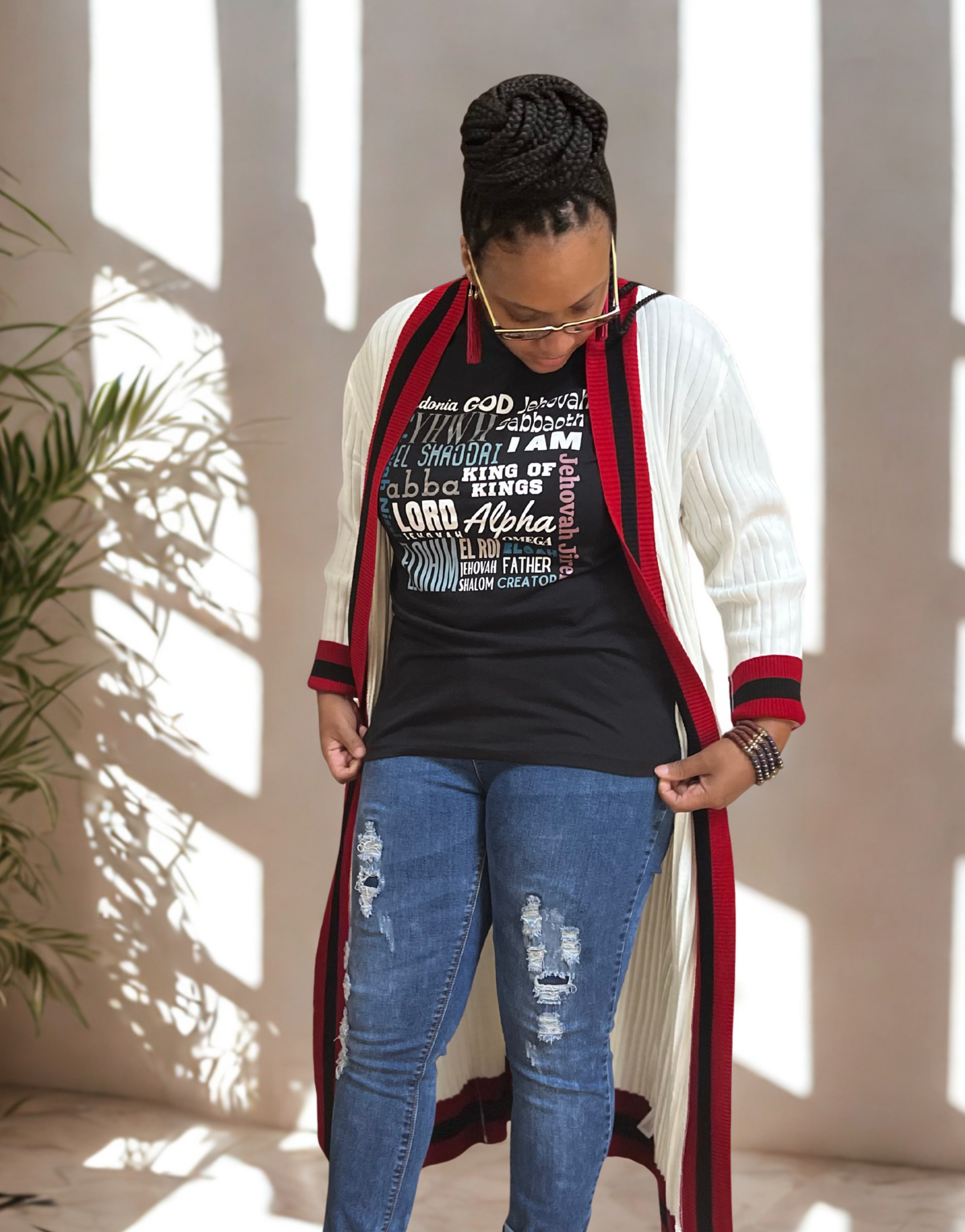 Woman wearing a long red and white cardigan over a black mission tee, I Am" graphic t-shirt and blue jeans, standing indoors with sunlight filtering through.