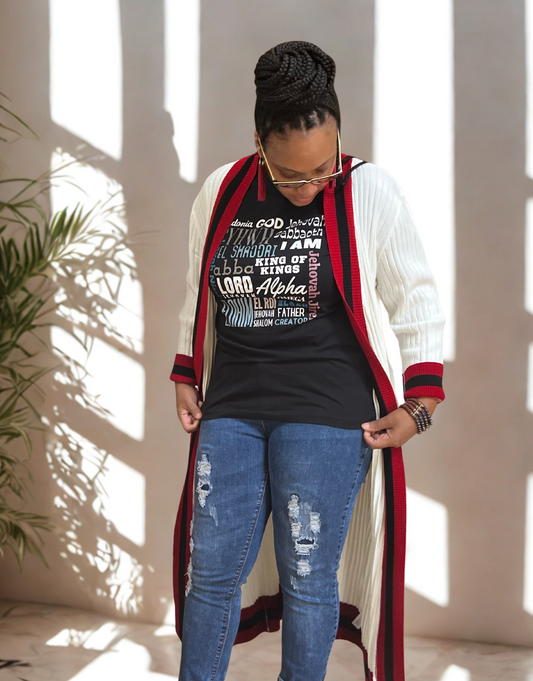 Woman wearing a long red and white cardigan over a black mission tee, I Am" graphic t-shirt and blue jeans, standing indoors with sunlight filtering through.